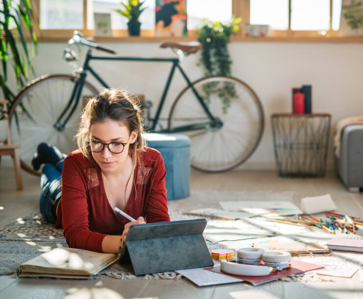 woman on the floor writing on a tablet device while looking at a book